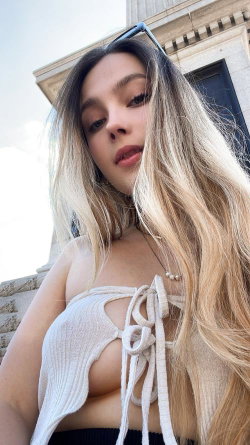 A woman with long ombre hair wears a white tied crop top, posing outdoors in front of a stone building.