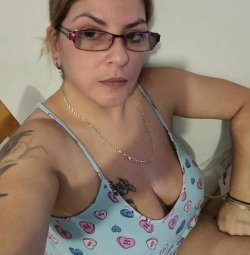 Woman with glasses and tattoos wearing a light blue heart-patterned tank top, posing indoors in a selfie-style shot.