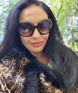 Woman with long dark hair wearing large sunglasses and a leopard-print fur coat outdoors, smiling softly.