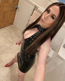 Brunette woman in a black polka-dot latex dress and red heels poses in a tiled bathroom, taking a selfie.