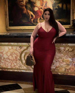 Curvy woman in a red mermaid gown poses confidently in an ornate room with a large classical painting behind her.