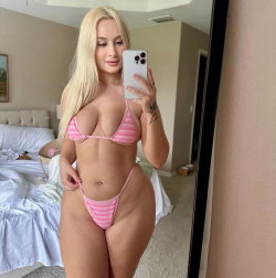 Blonde woman in pink striped bikini takes mirror selfie in bedroom, showing curvy figure and tattoo on arm.
