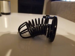 A black coiled spring object rests on a white table surface, with a blurred background featuring a metallic container