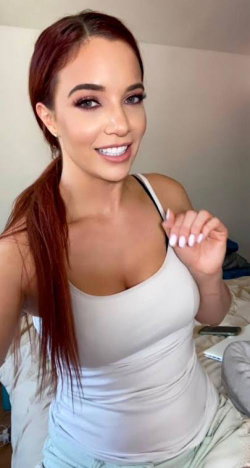 A woman with long red hair in a ponytail smiles while wearing a white tank top, sitting on a bed in a casual indoor