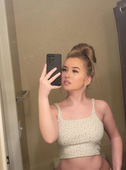 A blonde woman in a floral crop top takes a mirror selfie in a bathroom, hair styled in a bun.