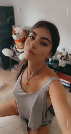 A woman with dark hair takes a selfie in a gray tank top, sitting indoors with stuffed animals and books in the