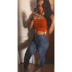Curvy woman with long dark hair takes a mirror selfie in an orange crop top and high-waisted jeans, standing indoors.