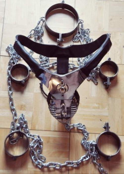 Metal and leather bondage gear with chains, cuffs, and a harness laid out on a wooden floor.