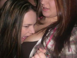 Two women close together, one with dark hair and a brown top, the other in a plaid shirt, in a dimly lit social setting.