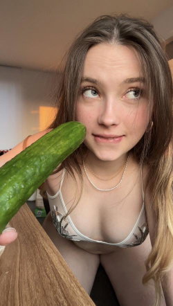 A young woman with long brown hair and a floral bra holds a cucumber near her chest in a close-up selfie indoors.