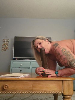 A woman with blonde hair and visible tattoos on her arm is topless, leaning over a wooden table while holding a large
