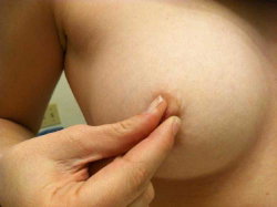 Close-up of a person's bare breast with hand gently touching the nipple area, skin tone light, indoor setting.
