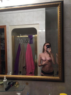 Blonde woman takes mirror selfie in bathroom, topless with black underwear, hand covering chest.