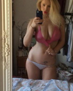 Blonde woman in pink bra and patterned thong takes mirror selfie in bedroom, hand on hip, visible tattoo on lower