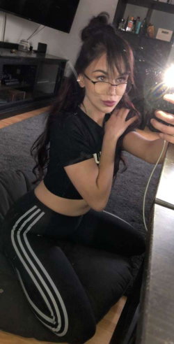 Brunette woman with glasses in black crop top and striped leggings, kneeling on a couch taking a mirror selfie in a
