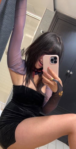 Brunette woman in a black velvet mini dress and sheer long sleeves takes a mirror selfie in a bathroom, one arm raised.