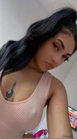 Brunette woman with long dark hair in a high ponytail, wearing a pink ribbed tank top, posing indoors with visible