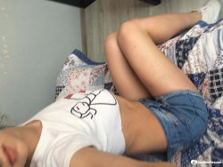 A woman lies on a patterned bed, wearing a white graphic tee and denim shorts, with her legs bent and arms partially