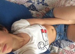 A woman lies on a patterned bed, wearing a white crop top and denim shorts, with her arm resting across her midsection.