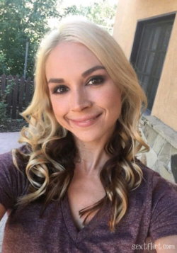 Blonde woman with wavy hair and makeup, wearing a purple V-neck top, smiling in a selfie outdoors near a building.