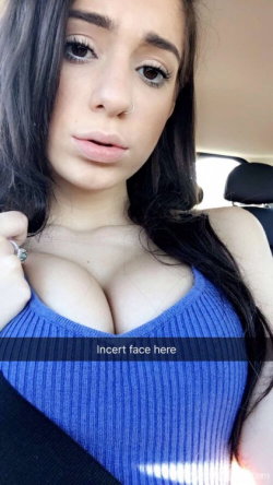Brunette woman with long dark hair wearing a blue ribbed top, taking a selfie inside a car with visible seatbelt.