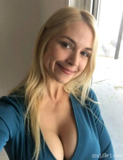 Blonde woman with long hair in a blue top, smiling at the camera in a selfie pose near a window.