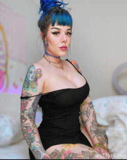 A woman with blue hair and full arm tattoos wears a black strapless top, sitting on a bed in a softly lit room.
