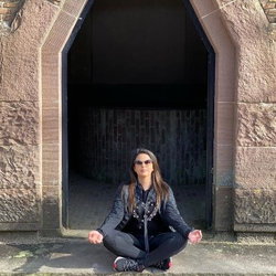 A woman with long dark hair sits cross-legged in front of a stone archway, wearing sunglasses, a black jacket, and