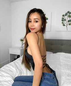 Asian woman with long dark hair in a black lace-up top and jeans, sitting on a bed in a bright bedroom.