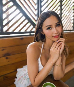 A woman with long dark hair and makeup wears a white spaghetti-strap top, sitting at a wooden table with a floral skirt