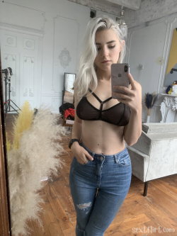 A blonde woman with long hair takes a mirror selfie in a black bra and ripped jeans, standing in a bright, decorated