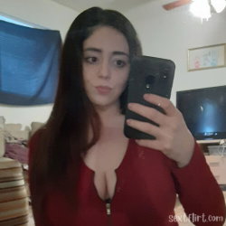 Brunette woman in a red zip-up top takes a mirror selfie in a bedroom setting.