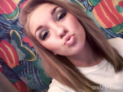 A young woman with long blonde hair and makeup makes a playful face in a selfie, lying on a colorful patterned blanket.