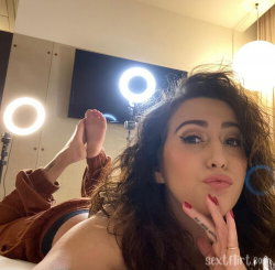 Brunette woman with wavy hair lies on bed, hand near face, wearing brown sweater and black underwear, ring lights in