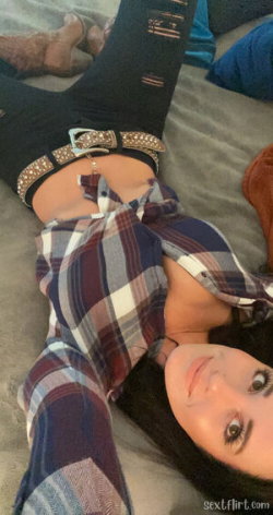 A woman with dark hair lies on a bed, wearing a plaid shirt and black ripped jeans with a studded belt, taking a selfie.