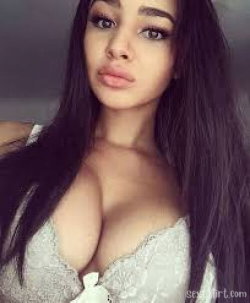 A woman with long dark hair and a curvy figure wears a white lace top, posing closely in a selfie-style shot indoors.