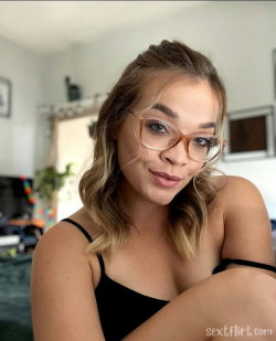 A woman with wavy blonde hair and glasses wears a black spaghetti strap top, sitting indoors in a casual home setting.