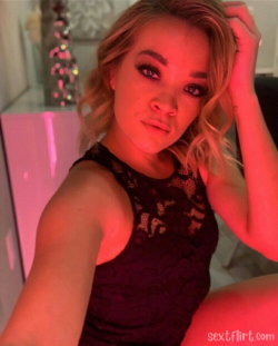 Blonde woman in a black lace top takes a selfie under red lighting, hand in hair, looking at the camera.