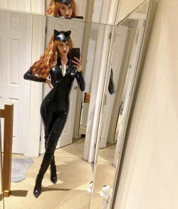 A woman with long red hair in a black catsuit and thigh-high boots takes a mirror selfie in a hallway.