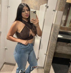 A curvy woman with long dark hair takes a mirror selfie in a brown halter top and ripped jeans, standing in a bathroom.