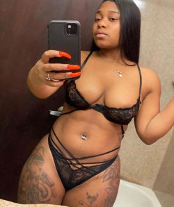 A Black woman with long dark hair takes a mirror selfie in black lace lingerie, showing tattoos on her thighs and