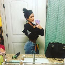 Brunette woman in a black cropped hoodie and jeans takes a mirror selfie in a bathroom, hair in a bun.