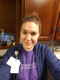 A woman with brown hair in a bun wears a purple shirt and blue jacket, smiling in a kitchen setting with wooden