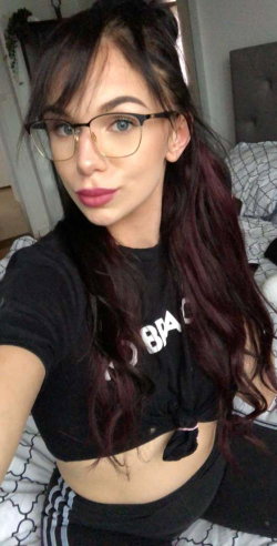 A woman with long dark hair and glasses takes a selfie in a bedroom, wearing a black crop top and striped pants.