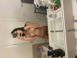 Brunette woman with curvy build takes mirror selfie in bathroom, topless and covering breasts with one hand.