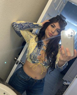 Brunette woman in a patterned crop top and jeans takes a mirror selfie, showing midriff and long hair.