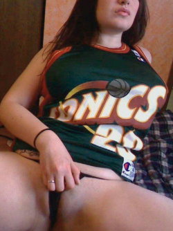 Woman with long dark hair wearing a green Sonics basketball jersey, sitting on a plaid surface with her legs slightly
