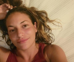 Woman with wavy brown hair lying on a bed, wearing a red tank top, smiling softly at the camera.