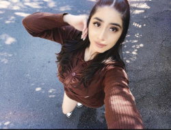 A young woman with long dark hair takes a selfie outdoors, wearing a brown ribbed top and black sneakers on a paved
