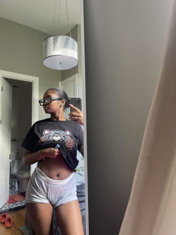 A woman with short braided hair and glasses takes a mirror selfie in a black graphic tee and white shorts, standing in